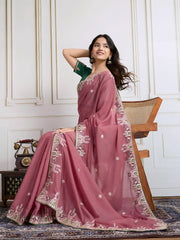  Woman wearing a dusty rose designer saree adorned with sequin detailing and an ornate embroidered floral border, paired with a contrasting green blouse embellished with small floral motifs. She stands elegantly in a classic white paneled room featuring a wooden armchair with a woven seat and a vintage rotary telephone on a carved wooden side table, all set atop an intricate patterned rug for a refined, traditional look.