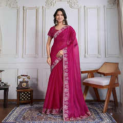 Woman wearing a magenta georgette saree adorned with subtle sequins and an ornate silver embroidered floral border, paired with a matching embroidered blouse. She stands elegantly in a classic white paneled room, accessorized with statement jhumka earrings and bangles, next to a vintage rotary telephone on a carved wooden side table and a modern wooden armchair set atop a decorative patterned rug for a graceful, timeless look.