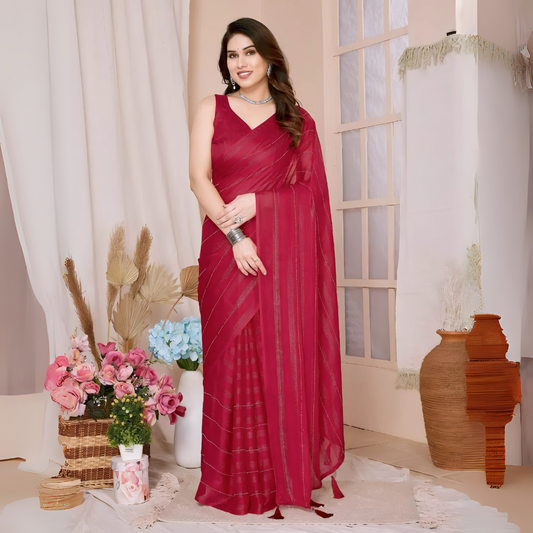 Woman wearing a black saree with subtle sequin stripes and a sleeveless blouse, accessorized with silver jewelry, standing in a modern interior with large windows, vases of pink and blue flowers, and neutral decor elements.