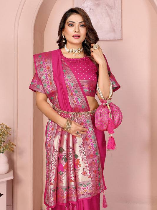 Woman wearing a magenta silk saree featuring a contrasting ethnic border with intricate geometric and floral patterns in gold and multi-color detailing, and matching tassels on the pallu. She pairs it with a matching blouse showcasing rich embroidery. She accessorizes with traditional choker, necklace, and bangles, standing elegantly in a pastel arched studio with potted plants and minimalist decor, presenting a refined and graceful ethnic look.