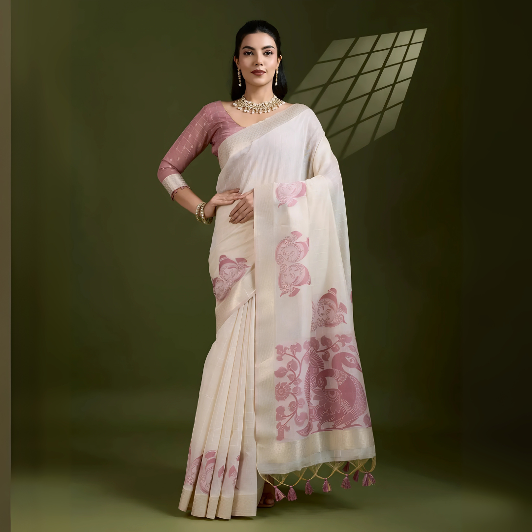 Woman wearing an ivory silk saree with pink floral and bird motifs, paired with a mauve blouse and pearl jewelry, posing in a contemporary studio with olive green walls and a windowpane shadow pattern in the background.