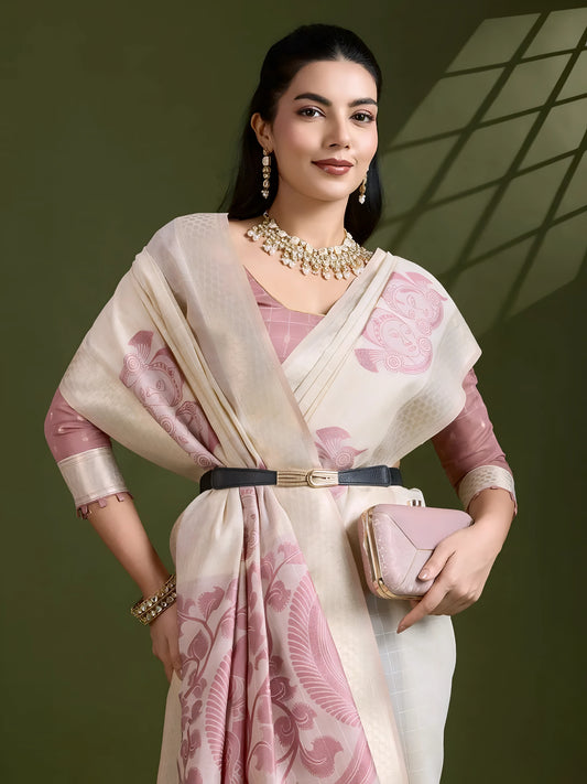 Woman wearing an ivory silk saree with pink floral and bird motifs, paired with a mauve blouse and pearl jewelry, posing in a contemporary studio with olive green walls and a windowpane shadow pattern in the background