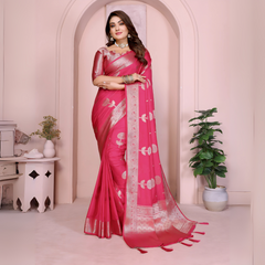 Woman wearing a magenta silk saree with silver zari border and floral motifs, paired with a matching blouse and silver jewelry. She poses gracefully in front of a pastel archway in an elegant studio setting, featuring a white table, decorative vases, and a potted green plant.