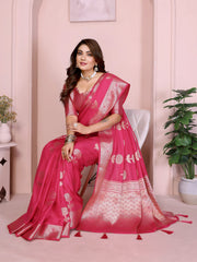 Woman wearing a magenta silk saree with silver zari border and floral motifs, paired with a matching blouse and silver jewelry. She poses gracefully in front of a pastel archway in an elegant studio setting, featuring a white table, decorative vases, and a potted green plant.