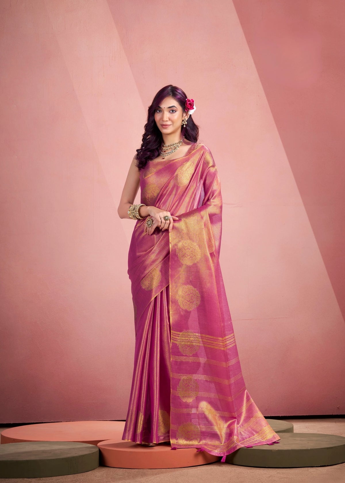 Woman in a pink and blue saree standing in an outdoor setting with stone walls and decorative elements.