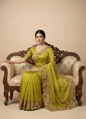 Woman in a green saree sitting on an ornate chair against a beige background