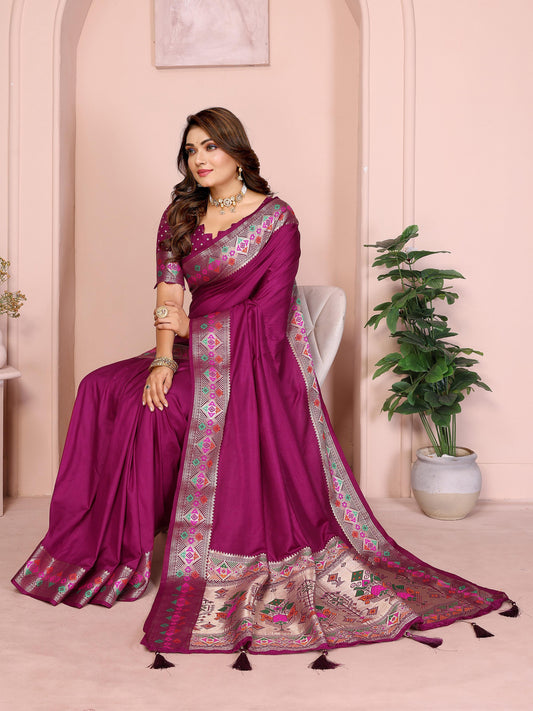 Woman wearing a plum purple silk saree with an ornate multicolor woven border featuring geometric and floral motifs, paired with a matching embroidered blouse and accessorized with a statement choker necklace, bangles, and rings. She poses gracefully in a pastel arched studio decorated with a white side table, floral vase, lush potted green plant, and a vintage-style chair, creating an elegant and sophisticated ambiance.