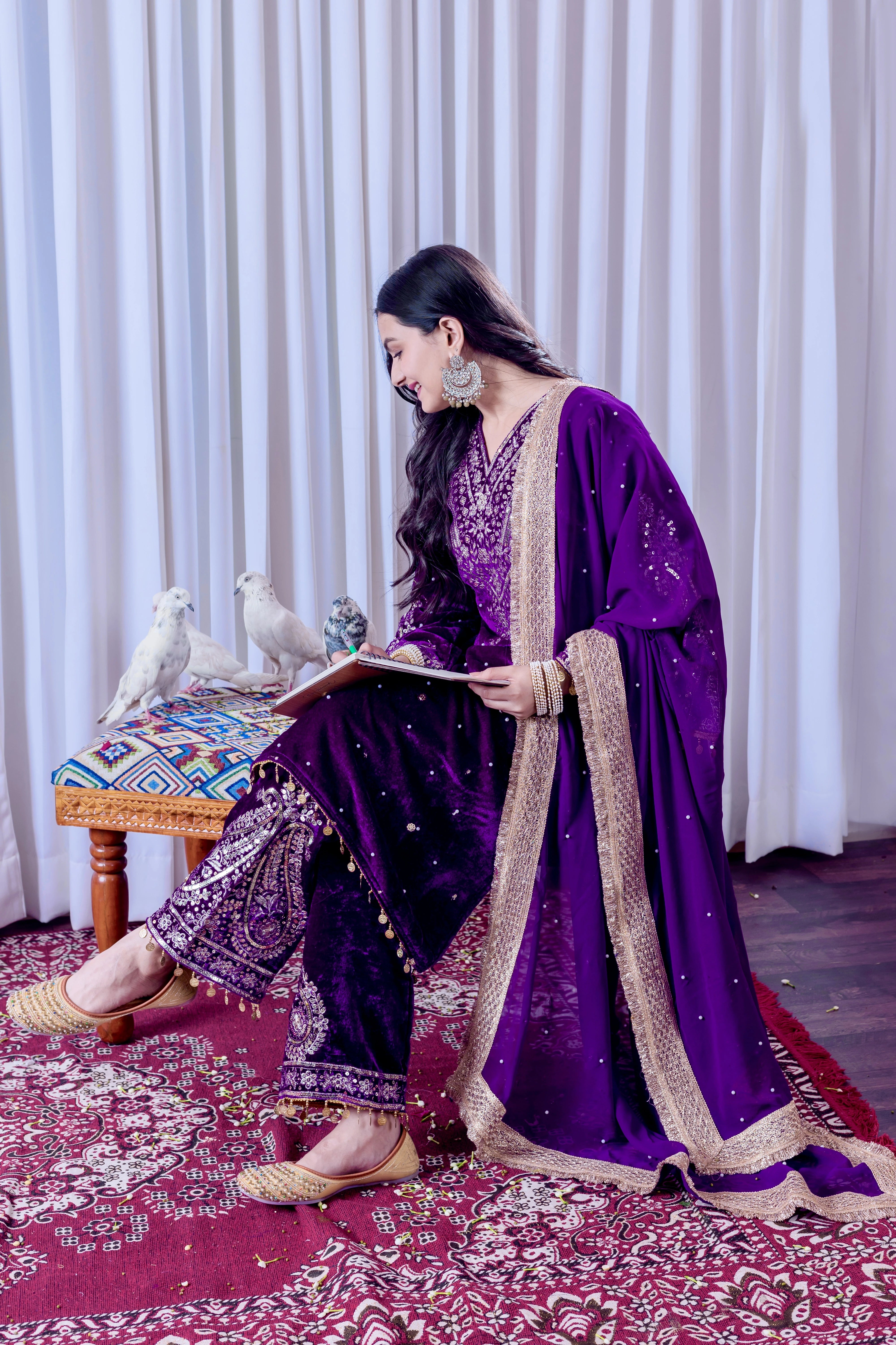 Woman in a purple traditional outfit sitting on a patterned rug with white curtains in the background.