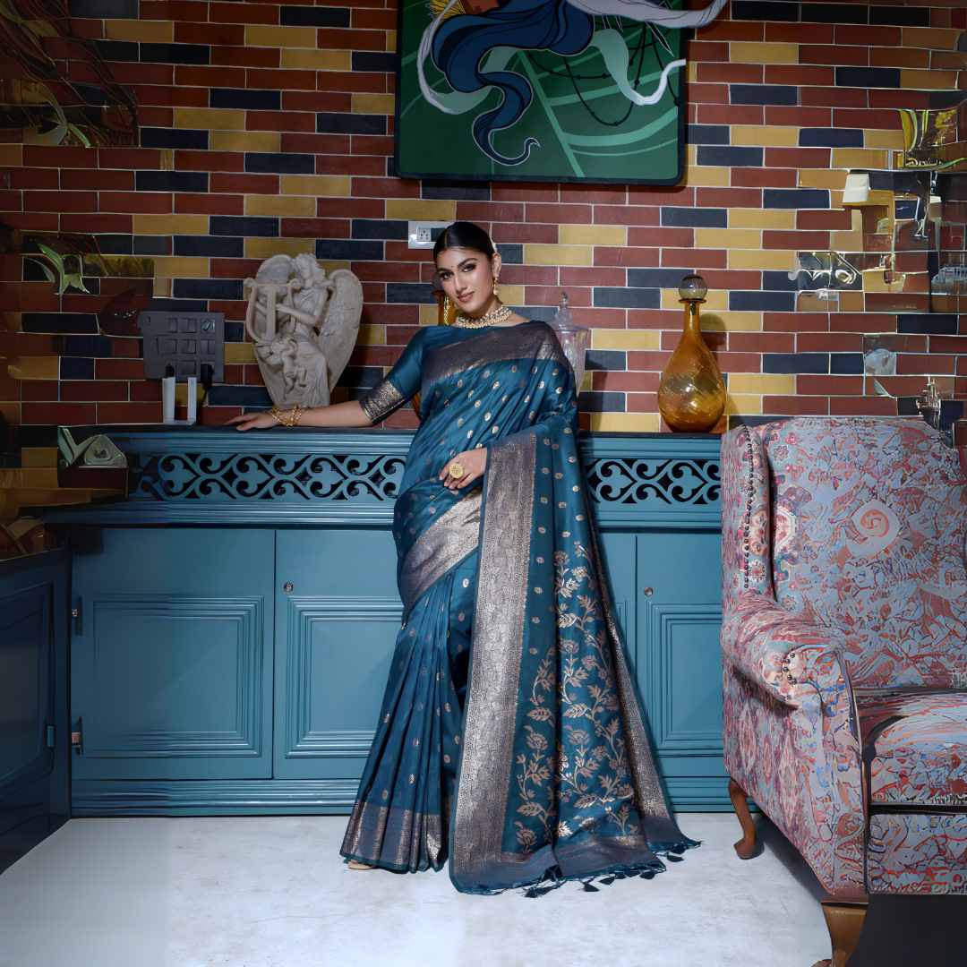 Woman wearing a teal silk saree with gold zari woven border and intricate floral patterns, accessorized with traditional jewelry and bangles, standing gracefully in a vintage-inspired room featuring a blue cabinet, multicolored brick wall, artistic decor, and an upholstered patterned armchair.