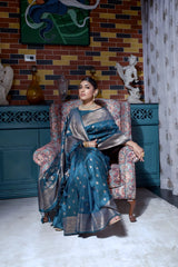 Woman wearing a teal silk saree with gold zari woven border and intricate floral patterns, accessorized with traditional jewelry and bangles, standing gracefully in a vintage-inspired room featuring a blue cabinet, multicolored brick wall, artistic decor, and an upholstered patterned armchair.