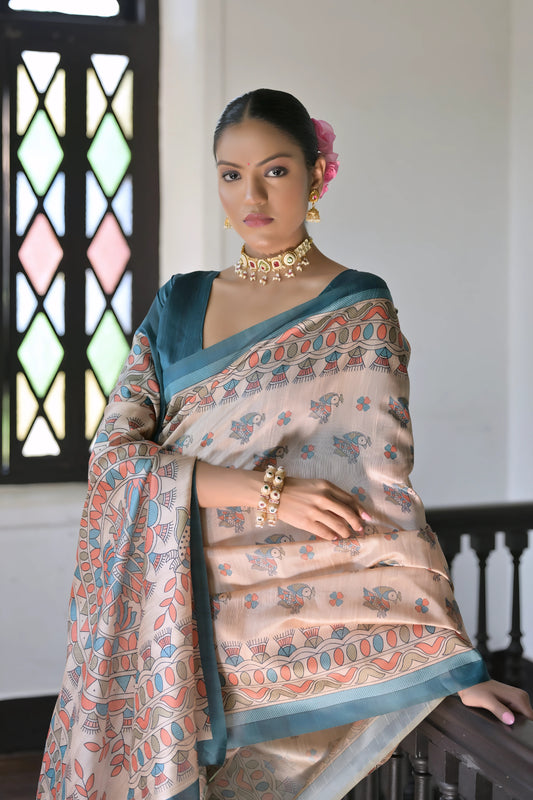Woman wearing a beige saree with intricate block print patterns and a blue border, paired with a blue blouse and floral hair accessory, posing in a sunlit vintage interior with wooden flooring, a cushioned bench, and a black railing near a window with stained glass panels.