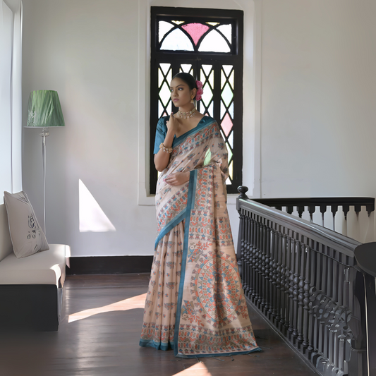 Woman wearing a beige saree with intricate block print patterns and a blue border, paired with a blue blouse and floral hair accessory, posing in a sunlit vintage interior with wooden flooring, a cushioned bench, and a black railing near a window with stained glass panels.