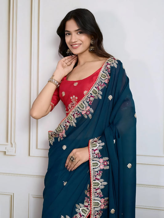 Woman wearing a teal blue designer saree embellished with beaded embroidery and ornate floral border, paired with a contrasting red blouse adorned with floral motifs. She poses stylishly in a classic white paneled room featuring a modern wooden armchair with a woven seat and a vintage rotary telephone on a carved side table, all set atop a patterned rug for an elegant, traditional look.