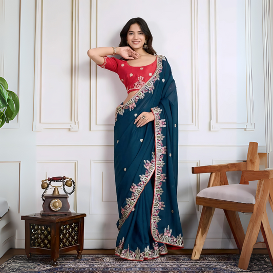 Woman wearing a teal blue designer saree embellished with beaded embroidery and ornate floral border, paired with a contrasting red blouse adorned with floral motifs. She poses stylishly in a classic white paneled room featuring a modern wooden armchair with a woven seat and a vintage rotary telephone on a carved side table, all set atop a patterned rug for an elegant, traditional look.