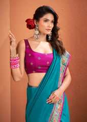 Woman wearing a purple blouse and teal saree with a red flower in her hair against a brown background