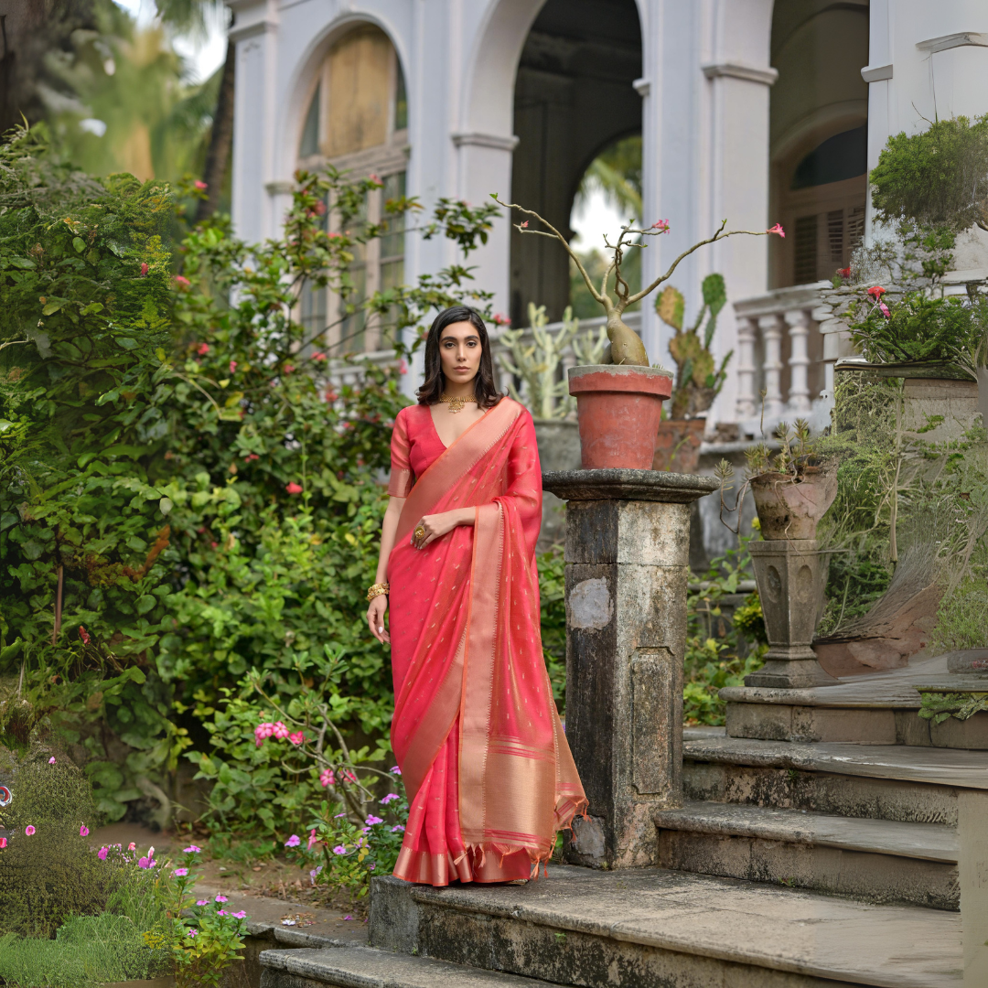 Woman wearing a coral silk saree with a gold border and subtle woven motifs, paired with a matching blouse and traditional jewelry, standing on the stone steps of a heritage mansion surrounded by lush greenery and potted plants.