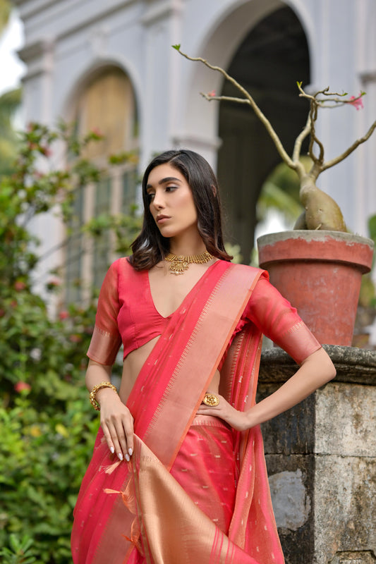 Woman wearing a coral silk saree with a gold border and subtle woven motifs, paired with a matching blouse and traditional jewelry, standing on the stone steps of a heritage mansion surrounded by lush greenery and potted plants.