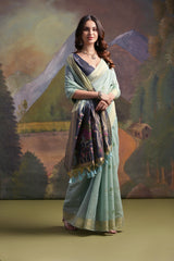 Woman wearing a pastel green silk saree with delicate floral embroidery and a contrasting navy blue blouse, standing gracefully in front of a painted mountain and forest backdrop, accessorized with traditional jewelry.