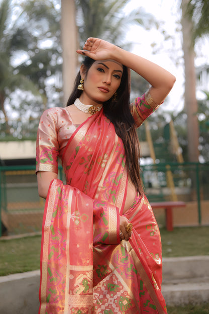 Woman wearing a coral pink Banarasi silk saree with gold zari motifs and a broad pallu featuring temple and floral patterns in green and gold, paired with a matching short sleeve blouse and gold jewelry. She stands on a manicured lawn in an outdoor garden setting with landscaped flower beds, fencing, and tall tropical trees in the background.