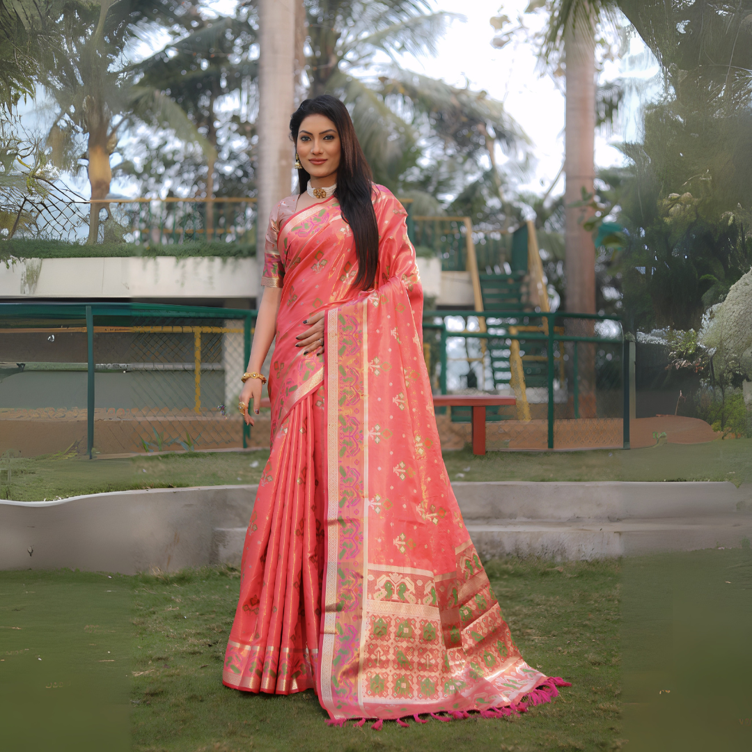 Woman wearing a coral pink Banarasi silk saree with gold zari motifs and a broad pallu featuring temple and floral patterns in green and gold, paired with a matching short sleeve blouse and gold jewelry. She stands on a manicured lawn in an outdoor garden setting with landscaped flower beds, fencing, and tall tropical trees in the background.