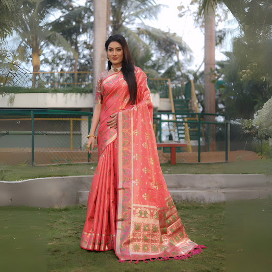 Woman wearing a coral pink Banarasi silk saree with gold zari motifs and a broad pallu featuring temple and floral patterns in green and gold, paired with a matching short sleeve blouse and gold jewelry. She stands on a manicured lawn in an outdoor garden setting with landscaped flower beds, fencing, and tall tropical trees in the background.