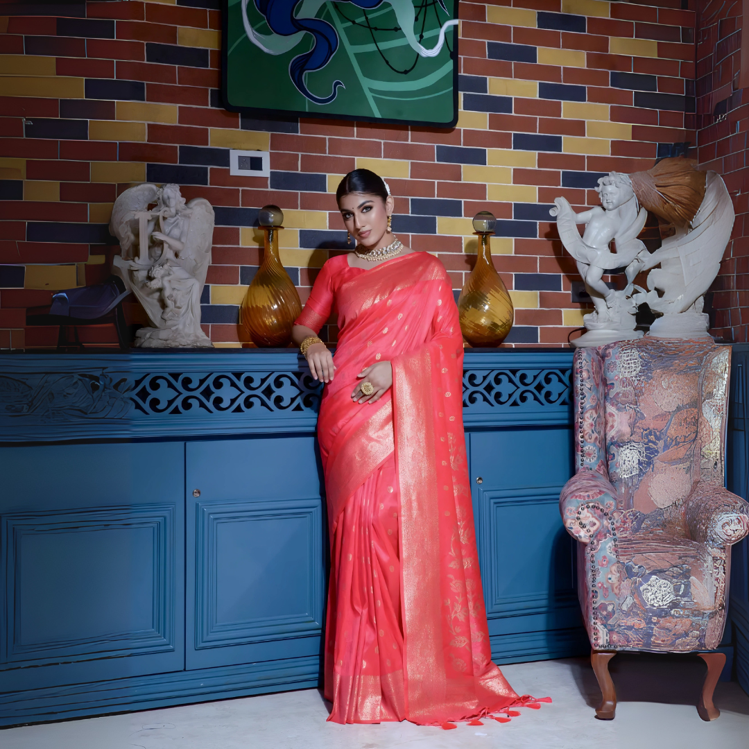 Woman wearing a coral pink silk saree with gold zari border and traditional polka dot motifs, accessorized with elegant gold jewelry, gracefully posing in a vintage-inspired room featuring a blue cabinet, colorful brick wall, decorative sculptures, and an upholstered patterned armchair.