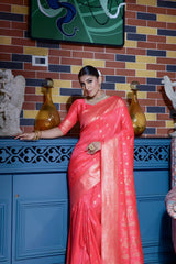 Woman wearing a coral pink silk saree with gold zari border and traditional polka dot motifs, accessorized with elegant gold jewelry, gracefully posing in a vintage-inspired room featuring a blue cabinet, colorful brick wall, decorative sculptures, and an upholstered patterned armchair.