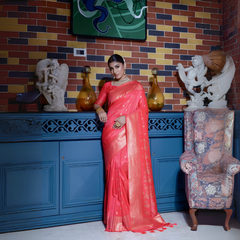 Woman wearing a coral pink silk saree with gold zari border and traditional polka dot motifs, accessorized with elegant gold jewelry, gracefully posing in a vintage-inspired room featuring a blue cabinet, colorful brick wall, decorative sculptures, and an upholstered patterned armchair.
