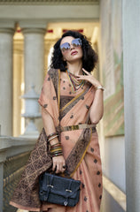 Woman in a peach saree with intricate patterns, holding a black handbag, in an elegant interior setting.