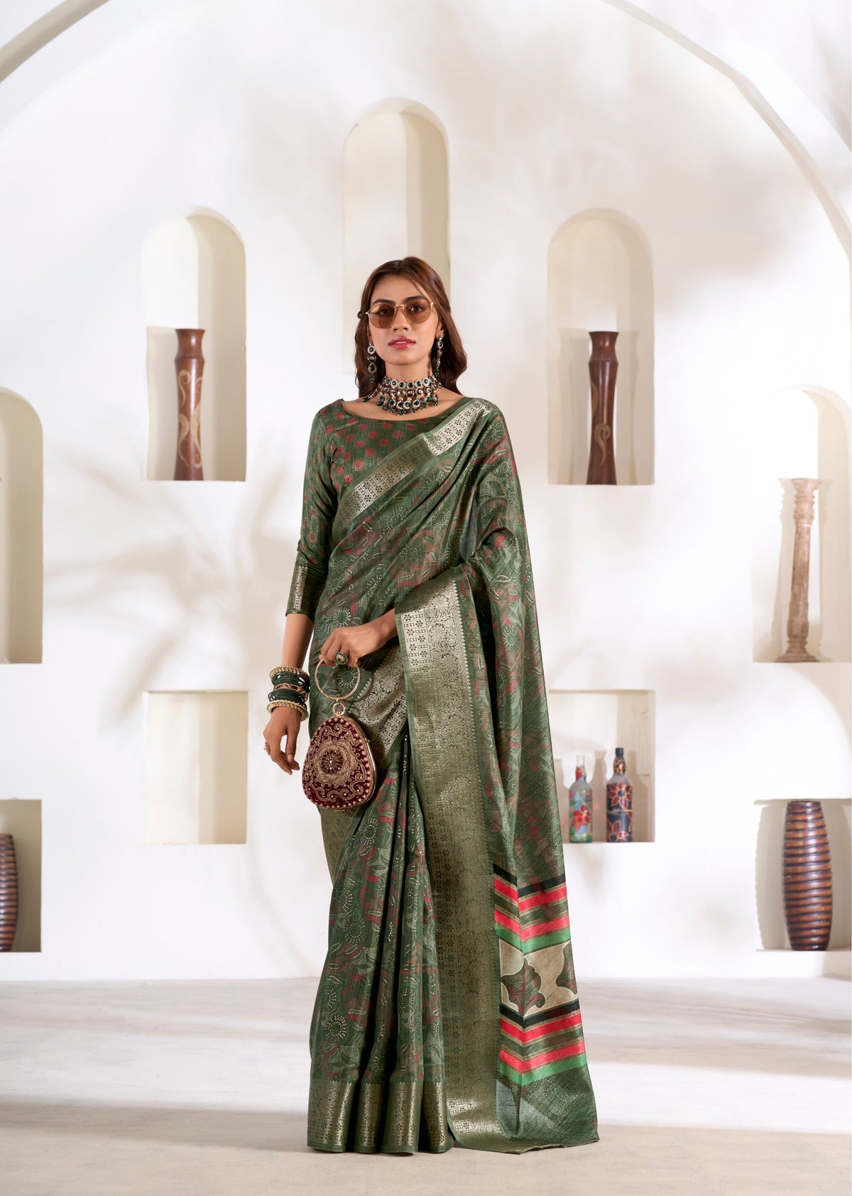 Woman in a green saree with red patterns standing in a white interior setting.