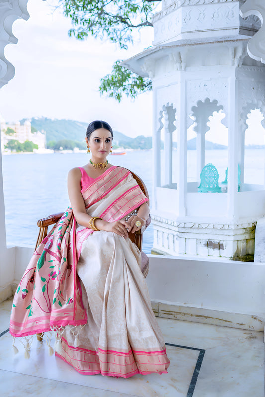 Woman wearing an ivory silk saree with pink border and handpainted floral pallu, styled with gold jewelry, posing in a traditional arched pavilion overlooking a serene lake and distant mountains.