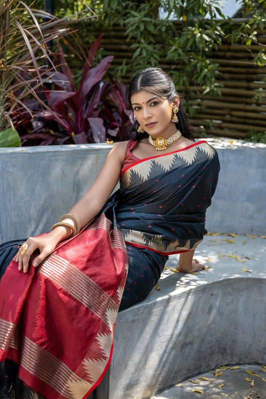 A woman models a dramatic Black Tussar Silk Saree with small red woven motifs. The saree has a beige temple-style border and a pallu featuring wide bands of red and beige weaving. She wears a red sleeveless blouse and a gold choker necklace.