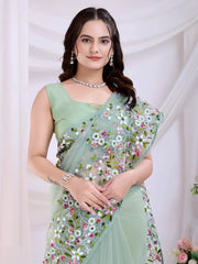 Woman wearing a mint green net saree with intricate multicolor floral embroidery and a matching sleeveless blouse, posing in a modern studio setting with white draped curtains and floral arrangements.