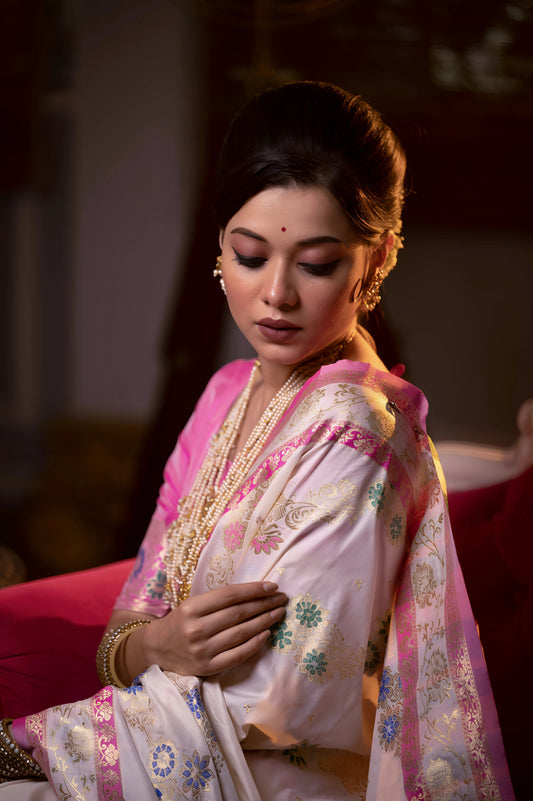  Woman wearing an ivory Banarasi silk saree with intricate pink floral zari border and pallu, paired with a pink blouse and layered gold jewelry, seated on a vintage white and red sofa in an elegant room with a gramophone, wooden blinds, and patterned flooring.

Related
File name: modern-minimalist-architecture
Alt text: Sleek modern minimalist architecture with clean lines and large glass windows
