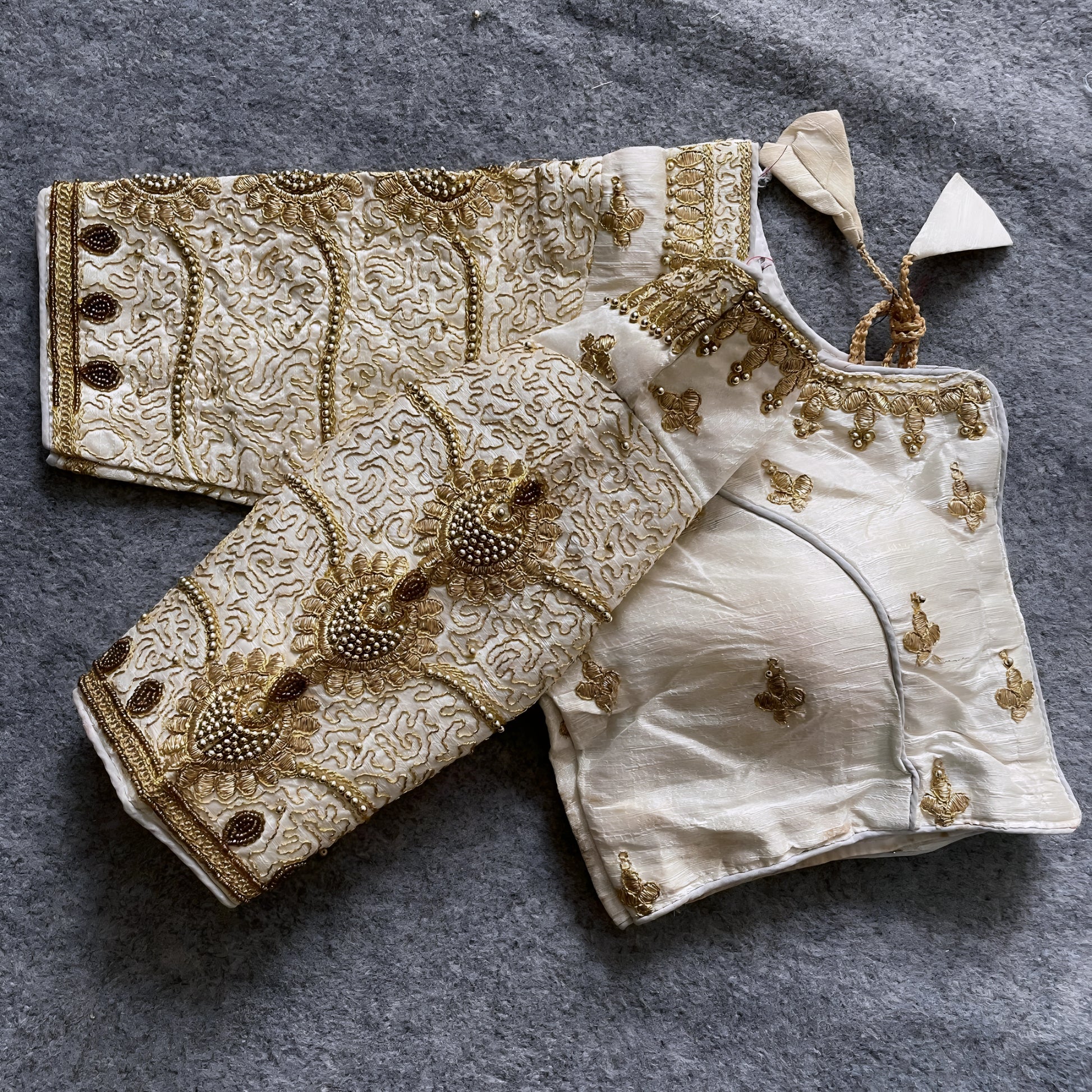 Traditional ivory silk blouse featuring intricate gold embroidery and beadwork with floral and paisley motifs on the sleeves and neckline, displayed flat on a textured surface with a bunch of artificial flowers and a woven mat in the background.