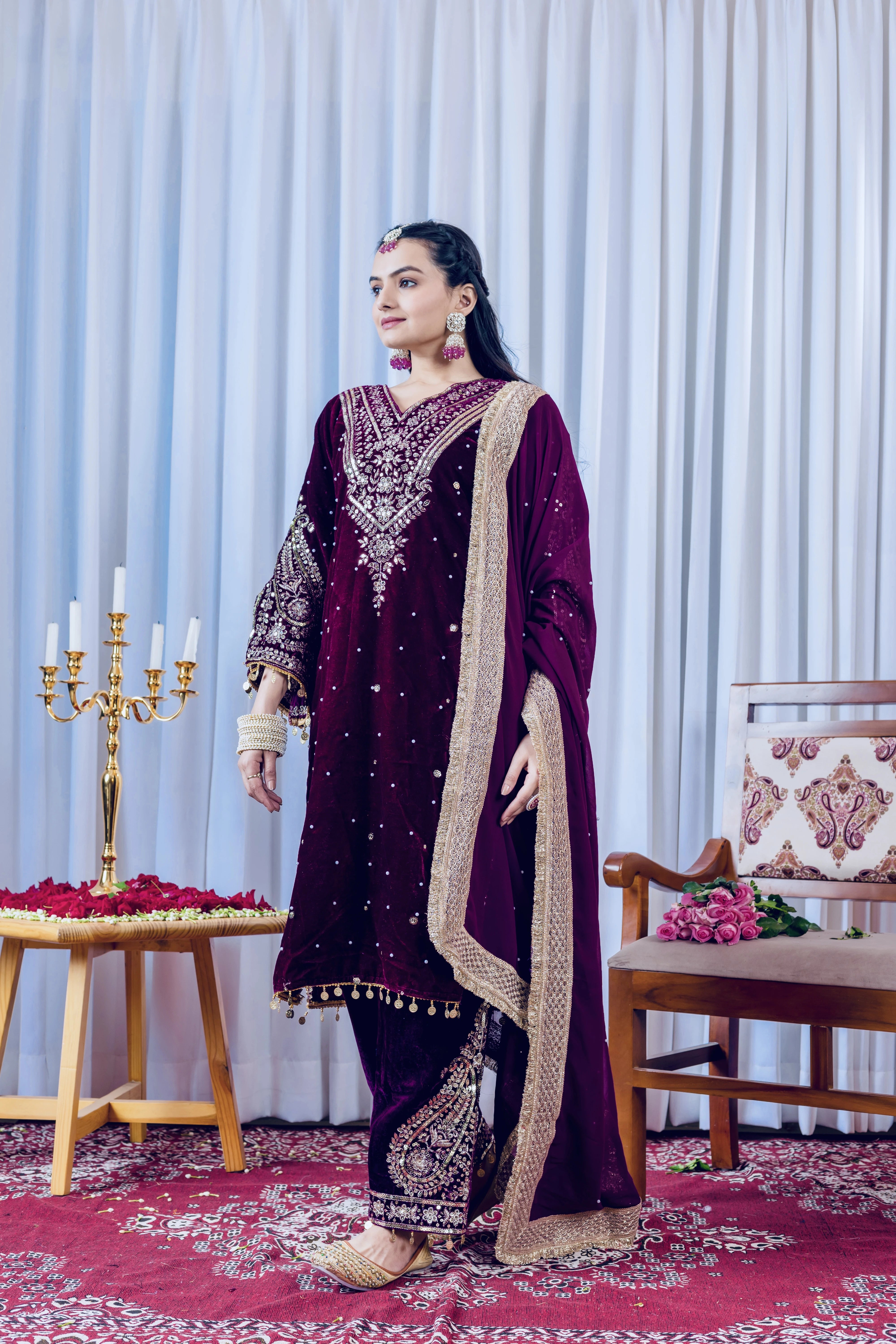 Woman in a dark purple embroidered traditional outfit standing in a decorated room with a candelabra and floral arrangements.