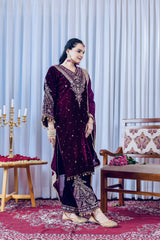 Woman in a dark purple embroidered traditional outfit standing in a decorated room with a white curtain and wooden furniture.