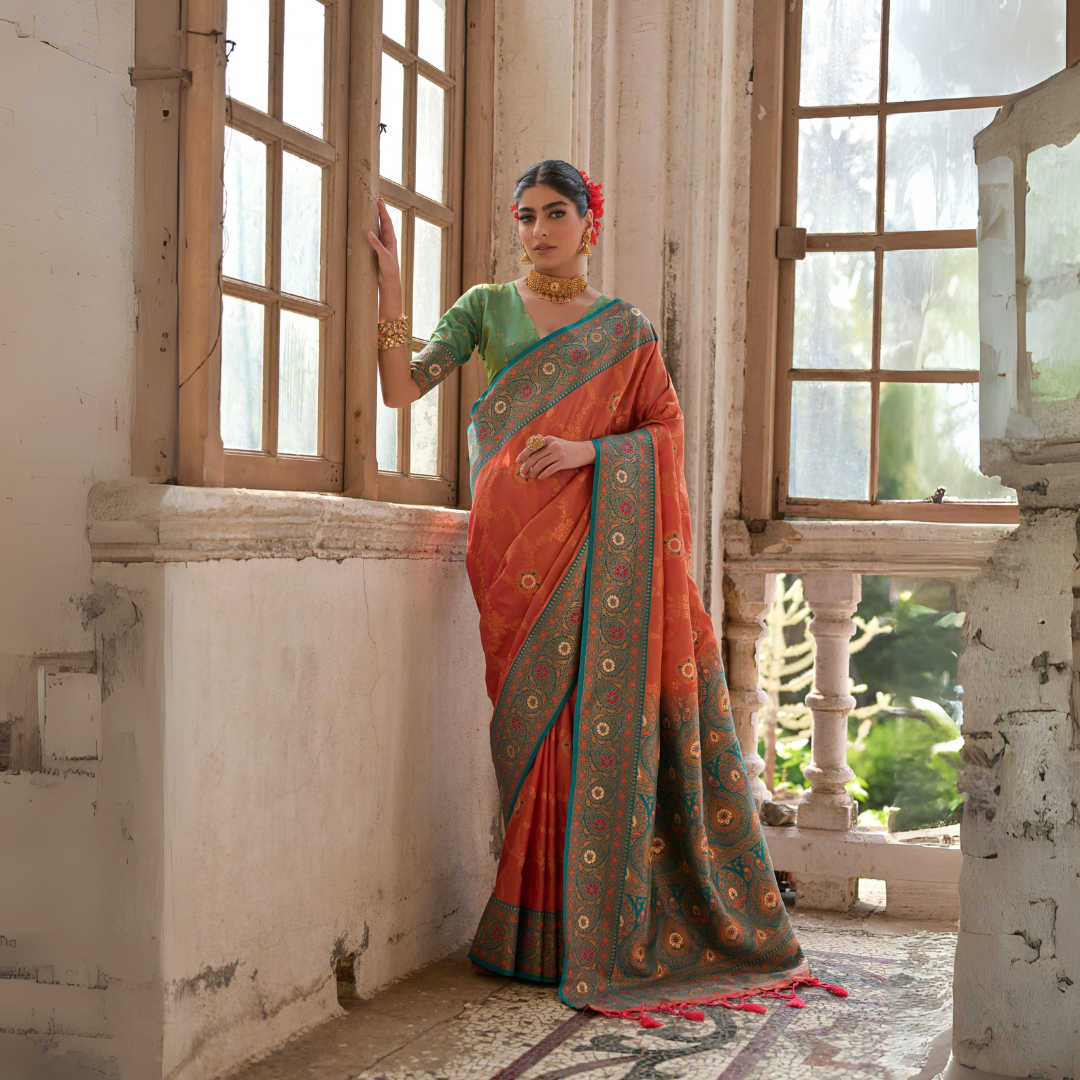 Woman wearing an orange silk saree with intricate gold and turquoise border, paired with a green blouse and traditional jewelry, standing by antique wooden windows in a vintage architectural setting with natural light streaming in.