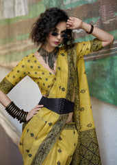 Woman wearing a yellow saree with black patterns against a textured wall.
