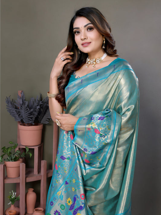Woman wearing an elegant aqua blue silk saree featuring a pastel sheen, intricate floral and polka dot motifs in multicolor threads, and a matching blue blouse with silverwork on the sleeves. She is accessorized with a choker necklace, bangles, and rings, posing gracefully in a contemporary studio with minimalist decor, including potted green plants and coordinated terracotta vases on wooden stands against a muted backdrop.
