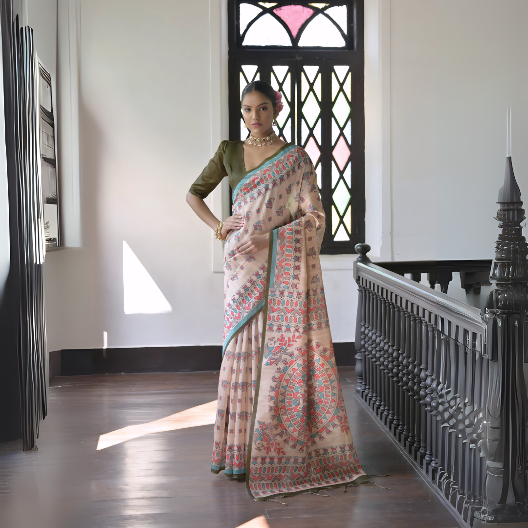Woman wearing a beige block print saree with teal and coral patterns and a green blouse, accessorized with a choker necklace and bangles, standing in a sunlit vintage interior with wooden flooring and a black window featuring stained glass panels.