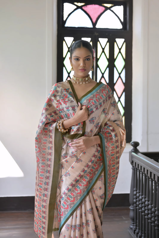 Woman wearing a beige block print saree with teal and coral patterns and a green blouse, accessorized with a choker necklace and bangles, standing in a sunlit vintage interior with wooden flooring and a black window featuring stained glass panels.