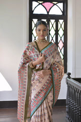 Woman wearing a beige block print saree with teal and coral patterns and a green blouse, accessorized with a choker necklace and bangles, standing in a sunlit vintage interior with wooden flooring and a black window featuring stained glass panels.