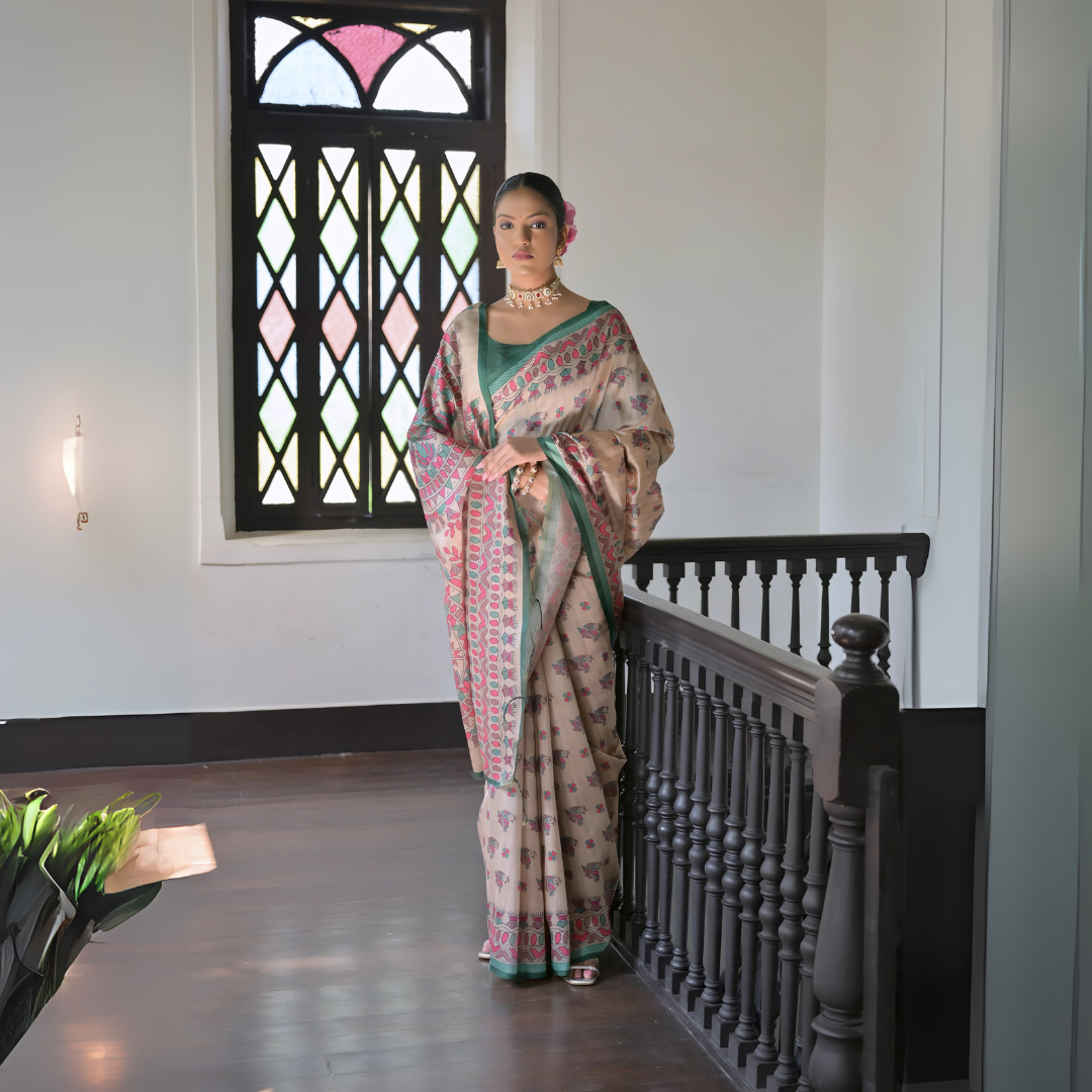 Woman wearing a beige block print saree with a green border and matching green blouse, accessorized with a choker necklace and floral hair accessory, standing in a sunlit vintage interior beside a stained glass window and dark wooden railing