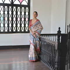 Woman wearing a beige saree with blue block print motifs and a pink border, paired with a deep pink blouse and pearl choker, standing beside a black wooden railing in a vintage interior with stained glass windows.