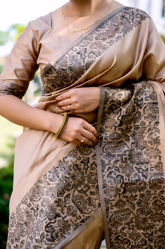 Woman wearing a beige silk saree with intricate brocade patterns and a matching blouse, standing gracefully on a stone pathway in a lush green garden with manicured lawns, trees, and sunlight filtering through the foliage.