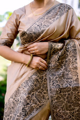 Woman wearing a beige silk saree with intricate brocade patterns and a matching blouse, standing gracefully on a stone pathway in a lush green garden with manicured lawns, trees, and sunlight filtering through the foliage.