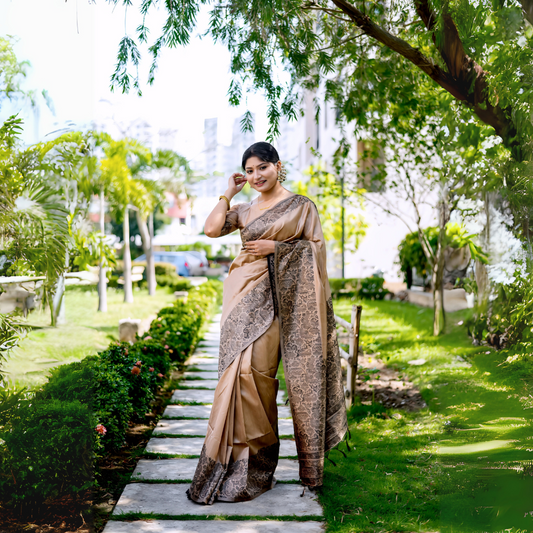 Woman wearing a beige silk saree with intricate brocade patterns and a matching blouse, standing gracefully on a stone pathway in a lush green garden with manicured lawns, trees, and sunlight filtering through the foliage.