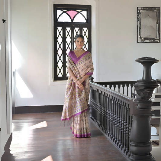 Woman wearing a beige silk saree with intricate floral motifs and a purple border, accessorized with a choker necklace and bangles, standing in a sunlit vintage interior with wooden flooring, black railing, and a window with stained glass panels.