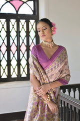 Woman wearing a beige silk saree with intricate floral motifs and a purple border, accessorized with a choker necklace and bangles, standing in a sunlit vintage interior with wooden flooring, black railing, and a window with stained glass panels.
